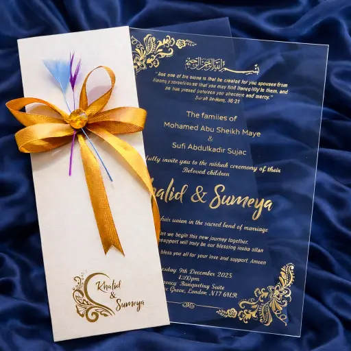Elegant wedding invitation suite on a wooden desk featuring deckle-edged paper with black script calligraphy, a translucent vellum wrap, and a gold botanical wax seal. Surrounding the cards are white roses, eucalyptus leaves, a gold pen, and an engagement ring in a velvet box